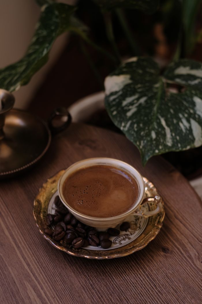 Services A close-up of a cup of coffee with beans on a decorative plate, set on a wooden table next to lush green foliage.