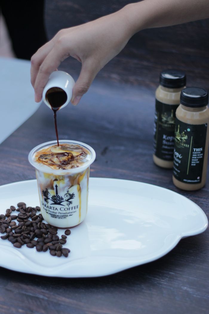 About A hand pours syrup into a Jakarta coffee cup surrounded by aromatic coffee beans on a tray.